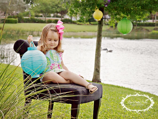 A young girl with a pink bow in her hair sits on a black chair by a lakeside, holding a blue paper lantern. She is wearing a colorful dress and sandals. In the background, there are hanging paper lanterns, green grass, trees, and a lake with ducks swimming.